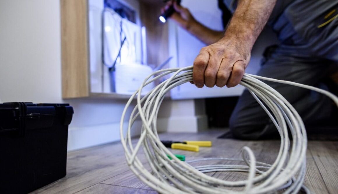 Close up shot of hand of aged electrician, repairman in uniform working, fixing, installing an ethernet cable in fuse box, holding flashlight and cable